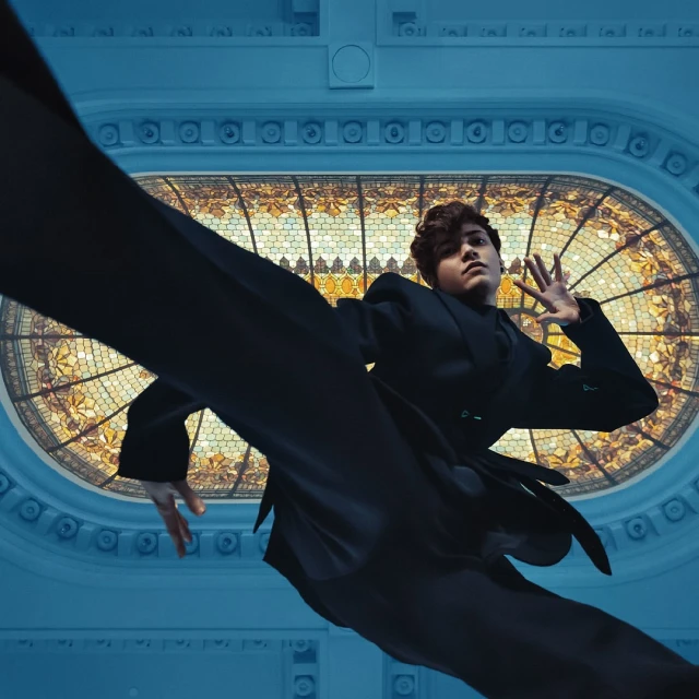 Fashionable young adult in black suit performs contemporary dance moves in front of a stained glass window and blue architectural details.