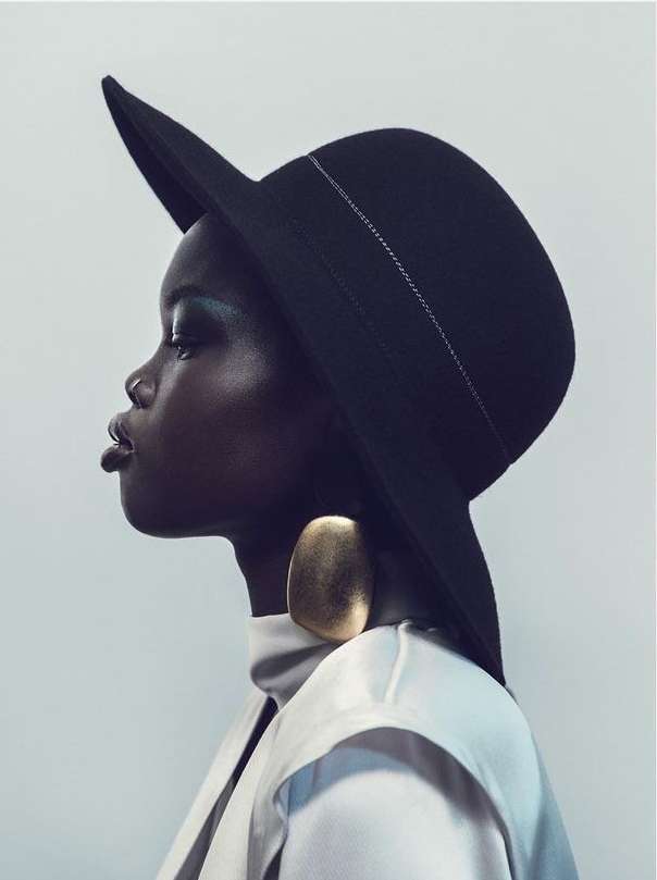 Profile of a dark-skinned woman wearing a large black hat and gold earring against a white background in a beauty fashion shot.