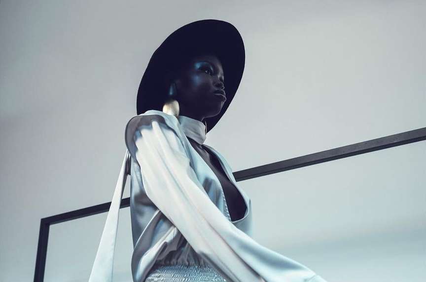 Fashionable African-American woman in a wide-brimmed hat and stylish top, looking up against a clean, minimalist backdrop.