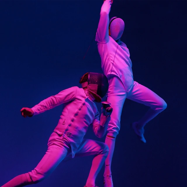 Two fencers in full gear engaged in a match, illuminated by neon pink and blue lights against a dark backdrop, creating a dramatic effect.