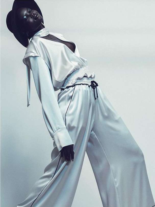 Fashion shot of a Black model in a satin pajama-style set and a wide-brim hat, posing against a neutral grey background in a studio.
