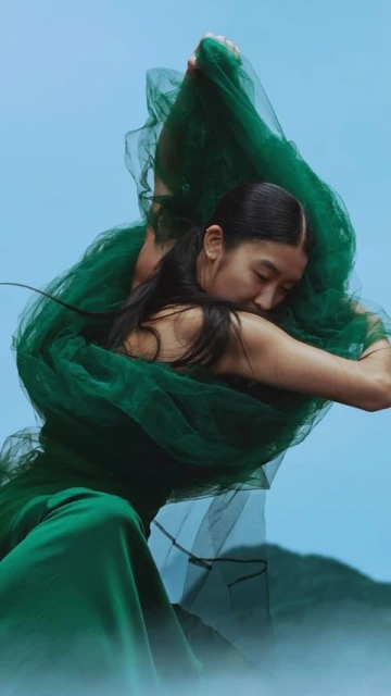 Woman in green dress and tulle fabric poses against a light blue background in a fashion editorial style studio shot.