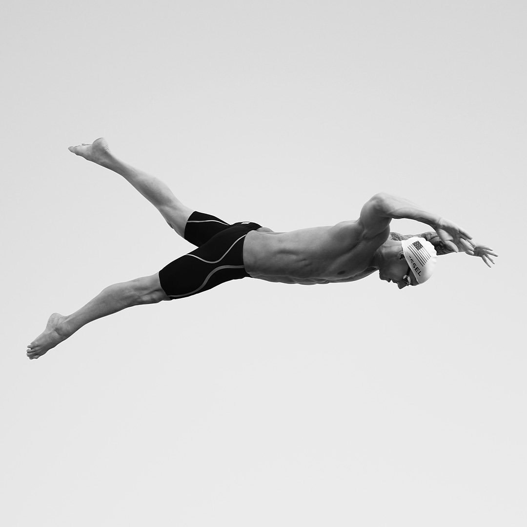 Monochrome action shot of a muscular swimmer with a swim cap performing the butterfly stroke against a light grey backdrop, arms outstretched.