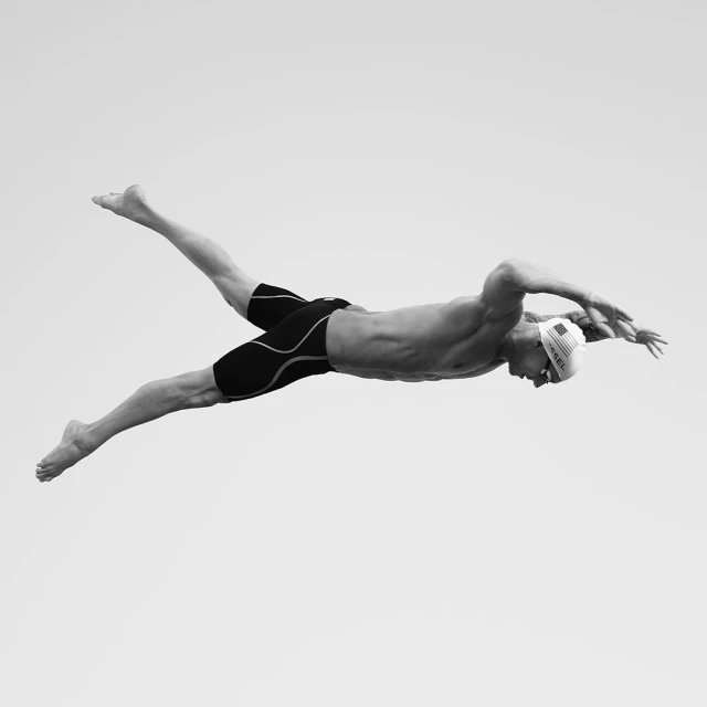 Monochrome action shot of a muscular swimmer with a swim cap performing the butterfly stroke against a light grey backdrop, arms outstretched.