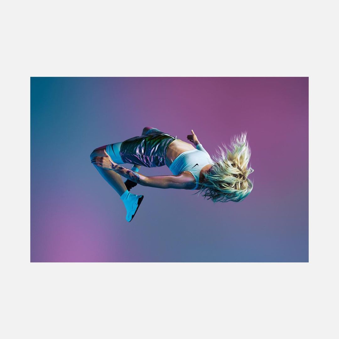 Athletic adult female with blonde hair performs backflip wearing sportswear against graduated pink and blue background in studio setting.