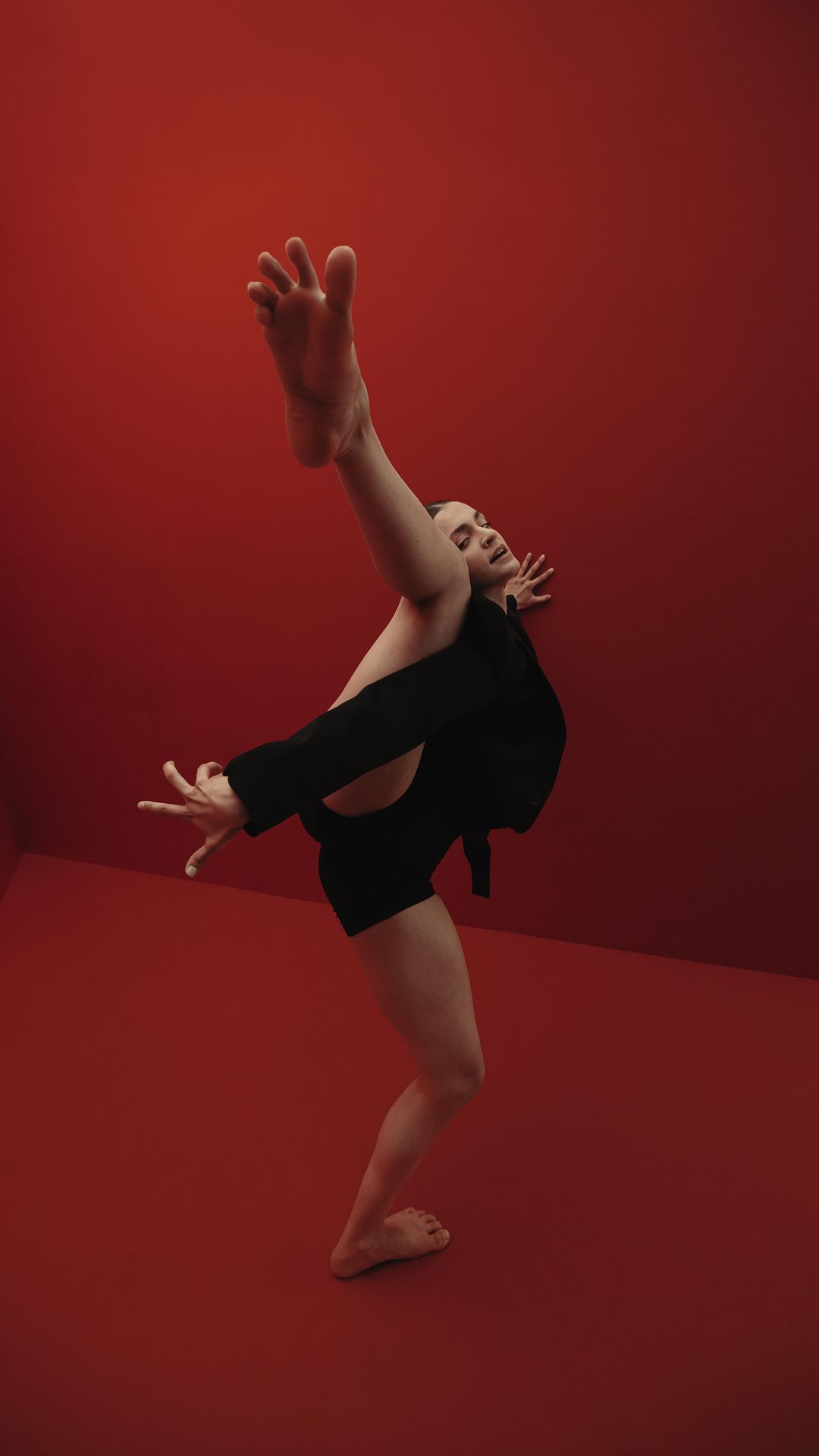 A person in a black jacket and shorts poses on a red background in a contorted position, leg extended toward the ceiling.