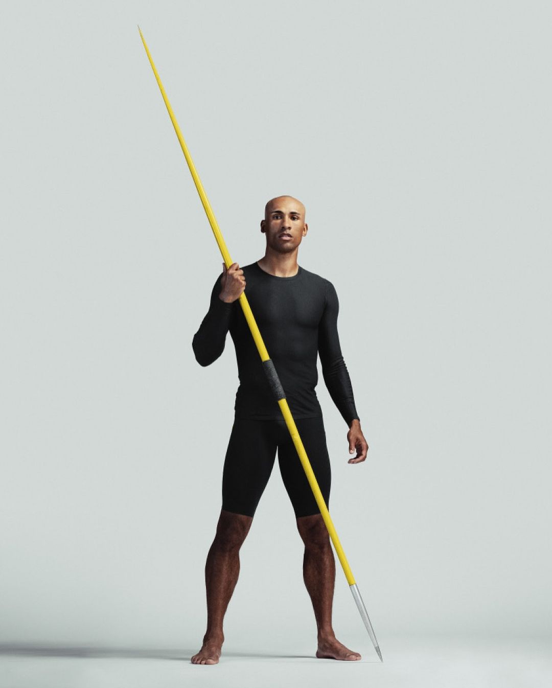 Athletic man in black activewear confidently poses in a full shot, holding a yellow javelin against a solid neutral background in a studio.