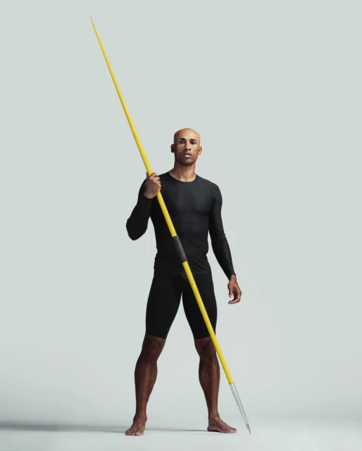 Athletic man in black activewear confidently poses in a full shot, holding a yellow javelin against a solid neutral background in a studio.