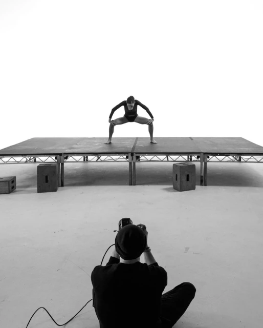 Monochrome shot of a woman in a bodysuit in a squat on a platform, photographed by a person with camera from a low angle indoors.