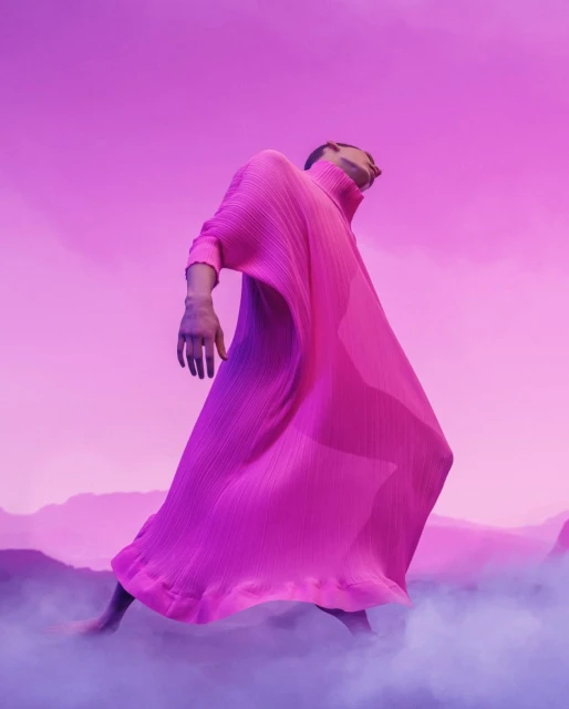 Person in long pink pleated dress striking a pose in front of pink sky and landscape with fog. Fashion editorial photography.