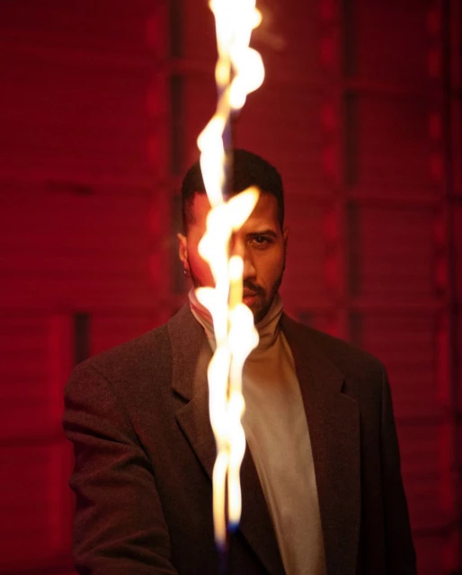 Portrait of adult male wearing a grey coat and beige turtleneck sweater, standing indoors with flames and a red wall in the background.