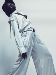 Black woman wearing a wide-brim hat and white satin pajama-style outfit posing dramatically against a white studio background.