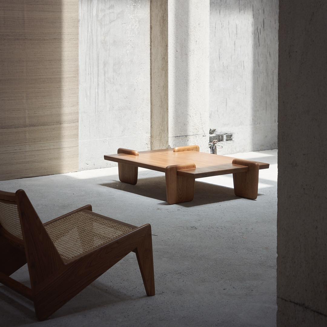 Minimalist interior featuring a light wood coffee table and chair, set against a concrete wall in a sunlit space.