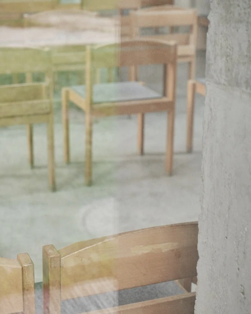 Several light brown wooden chairs with light grey seats are reflected in a shiny window or glass partition indoors.