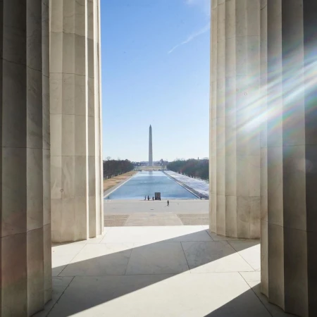 View of the Washington Monument through the columns of the Lincoln Memorial on a clear day, reflecting in the reflecting pool.