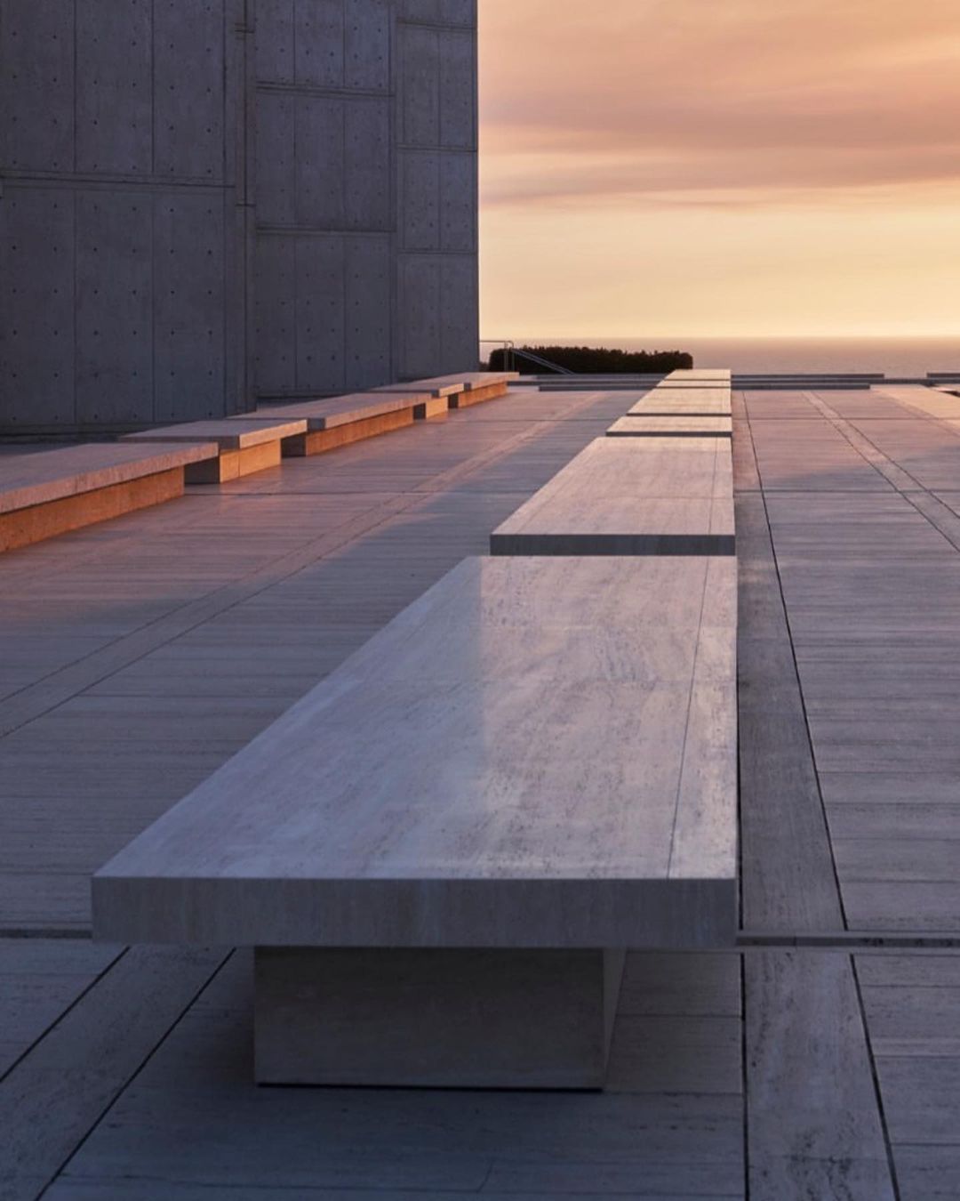 Architectural view of a stone bench on a paved outdoor terrace, with a concrete building and a sunset over the ocean in the background.