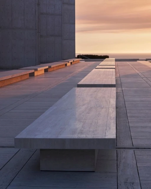 Architectural view of a stone bench on a paved outdoor terrace, with a concrete building and a sunset over the ocean in the background.