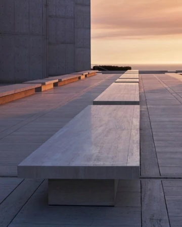 Architectural view of a stone bench on a paved outdoor terrace, with a concrete building and a sunset over the ocean in the background.