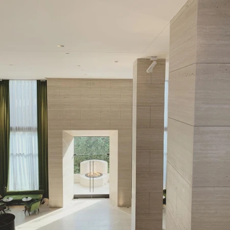 Open-plan living room featuring travertine walls, light wood flooring, and a view to a modern outdoor fireplace and patio area.