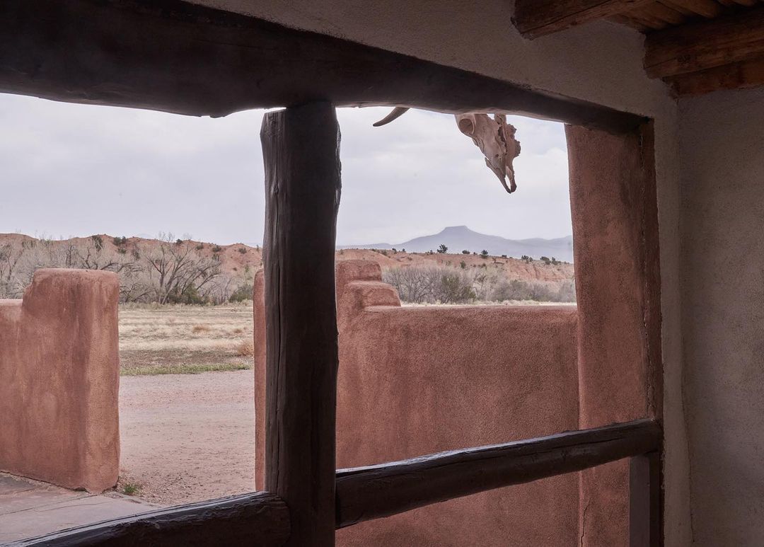 Adobe home with wooden beams frames a view of a mountain in the distance and a cow skull hanging above the doorway.