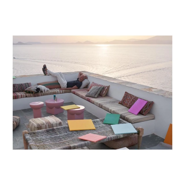 Adult male relaxing on a cushioned bench with a dog in an outdoor lounge area, showcasing a scenic view of the ocean.