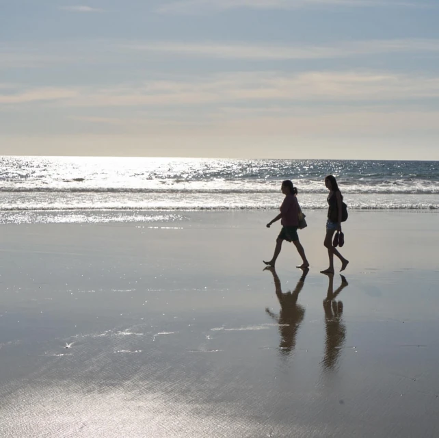 Two women silhouetted walking barefoot on a wet sandy beach near the ocean under a bright sunny sky with some clouds reflecting.