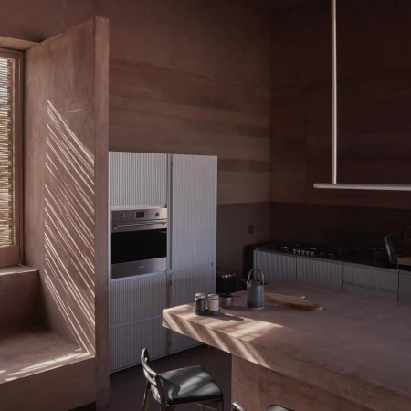 Interior view of a modern kitchen with light wood cabinets, stainless steel appliances, and a dark countertop with chairs in foreground.
