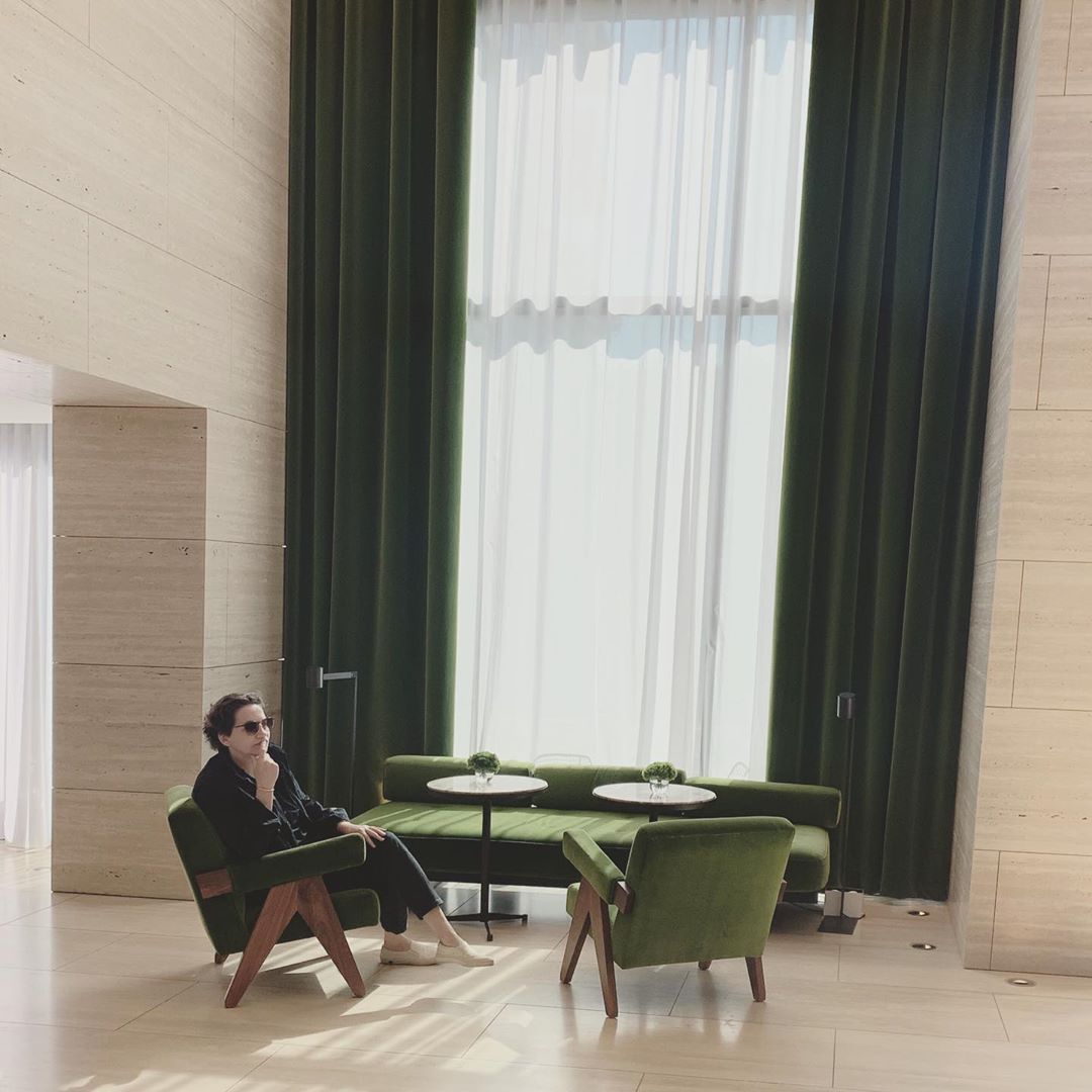 Adult male in sunglasses sits pensively in a green chair in a modern lobby with high ceilings, light stone walls, and dark green drapes.