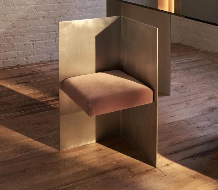 A modern chair with a geometric design featuring a velvet cushion, set on a wooden floor against a painted brick wall. 