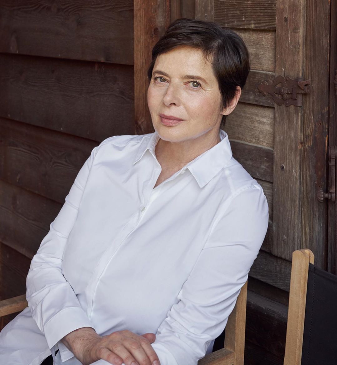 Portrait of an adult woman with short dark hair wearing a white shirt, seated outdoors in front of weathered wood panels.