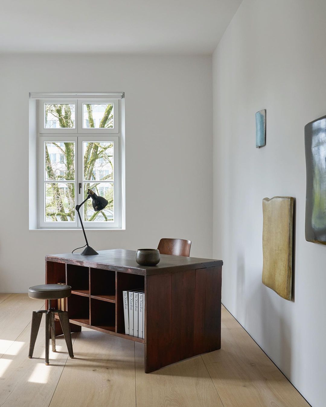 Desk with books and lamp in bright room with minimal decor, artworks on wall, natural light from window, light wood floor.
