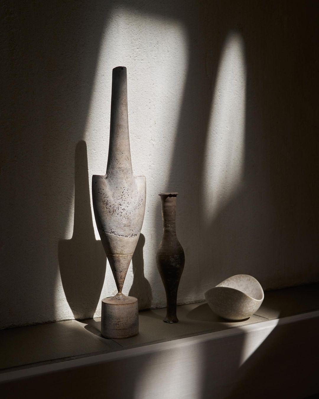 Still life of beige sculptural vases in a shaft of sunlight, arranged on a ledge against a pale beige wall, modern art.