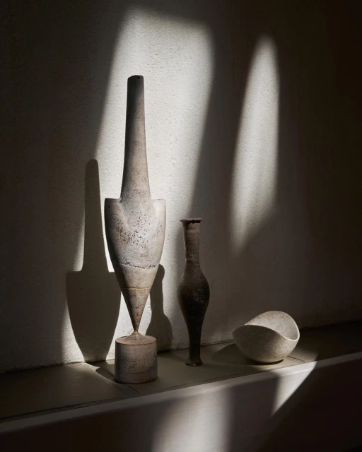 Still life of beige sculptural vases in a shaft of sunlight, arranged on a ledge against a pale beige wall, modern art.