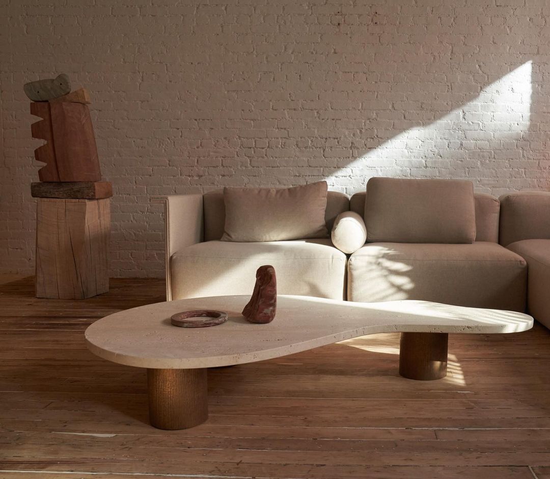 Modern living room interior featuring a travertine coffee table, beige sectional sofa, and brick wall with natural light.