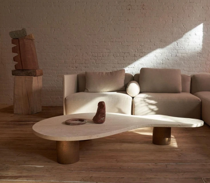 Modern living room interior featuring a travertine coffee table, beige sectional sofa, and brick wall with natural light.