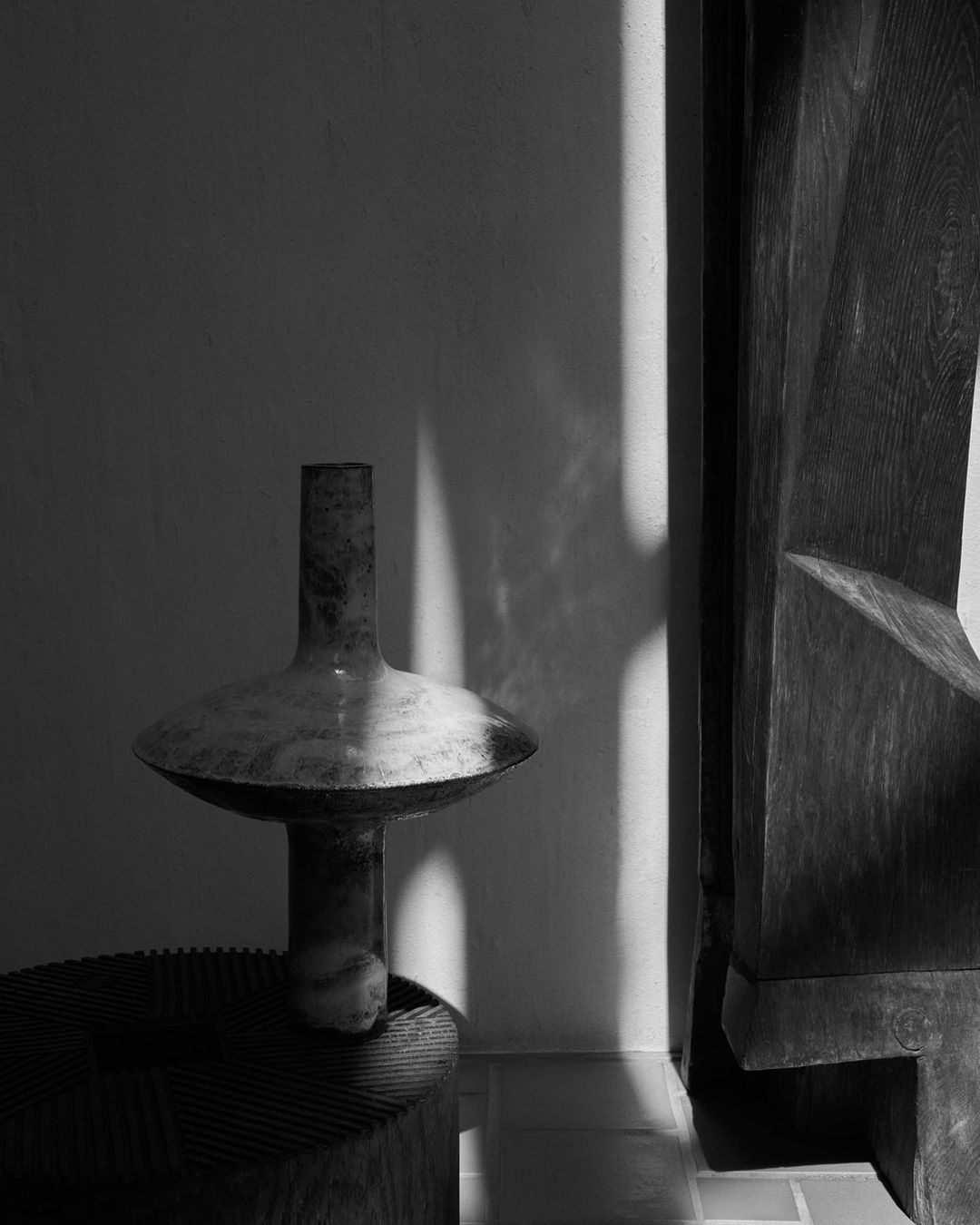 Monochrome still life featuring a sculptural ceramic vessel on a wooden base beside a carved wooden sculpture in a room with soft light.