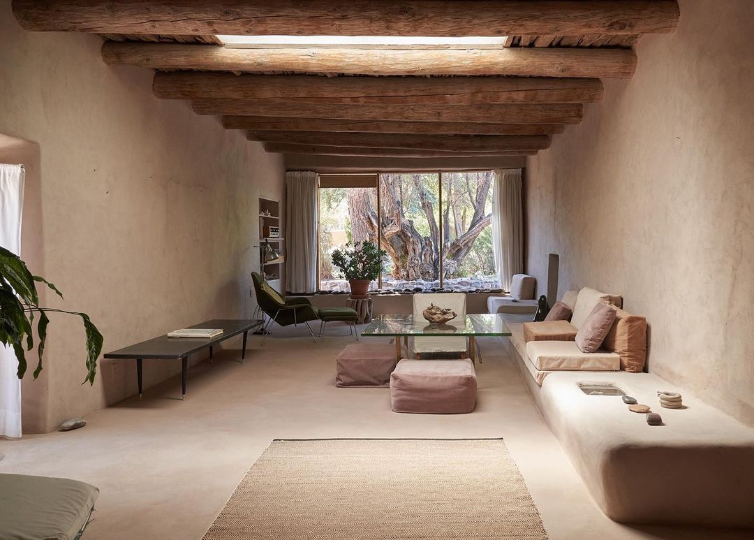Living room interior with earthen walls, wood-beamed ceiling, large window view of trees, sofa, chairs, glass table, rug and plants.