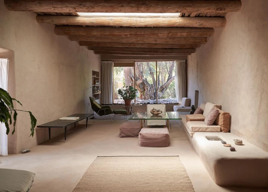 Living room interior with earthen walls, wood-beamed ceiling, large window view of trees, sofa, chairs, glass table, rug and plants.