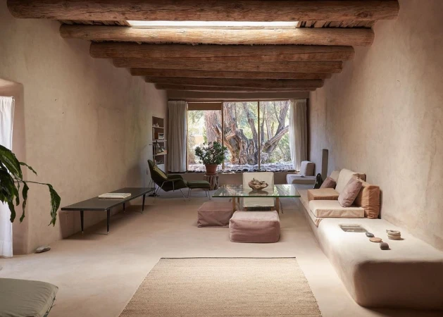 Living room interior with earthen walls, wood-beamed ceiling, large window view of trees, sofa, chairs, glass table, rug and plants.