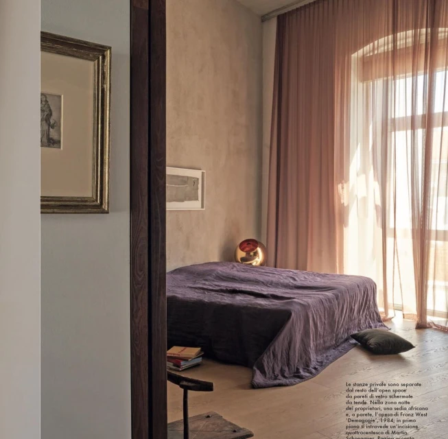 Bedroom interior with a made bed in front of mauve sheer curtains and a golden sphere lamp, illuminated by window light.