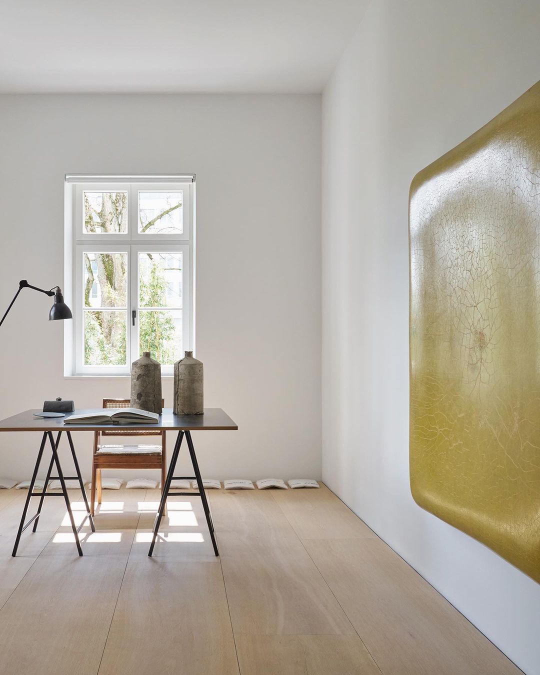 Minimalist interior with vases on a black table, wooden floors, and a modern gold-colored wall art piece in a white-walled room.