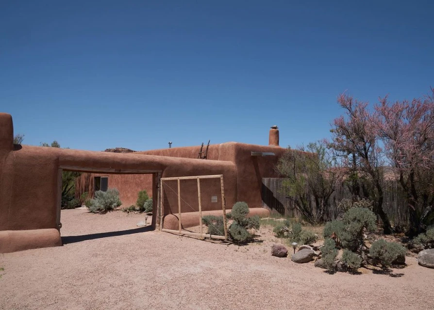 Adobe-style home with stucco walls, a chimney, and an arched gate, set against a clear blue sky in a desert landscape with rocks and trees.