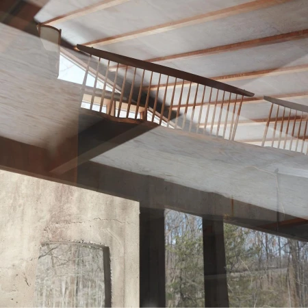 Interior view shows a staircase with a brown wooden railing and a concrete wall under a roof with wooden beams; sunlight streams through.