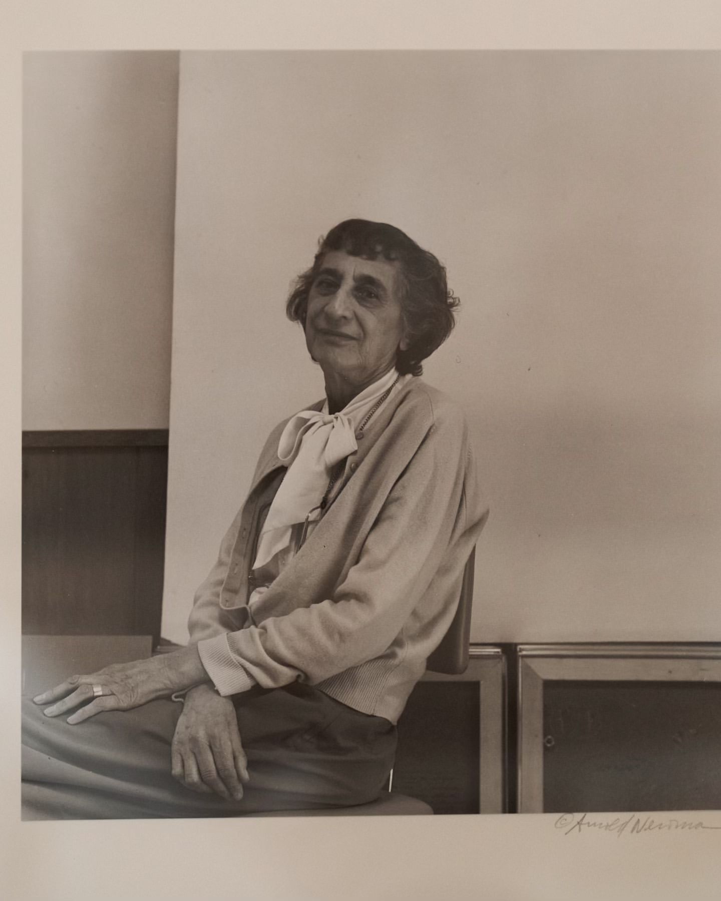 Portrait of an elderly woman with short, dark hair, wearing a light cardigan and scarf, seated indoors against a neutral background.