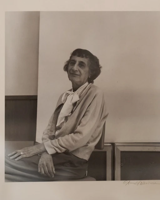 Portrait of an elderly woman with short, dark hair, wearing a light cardigan and scarf, seated indoors against a neutral background.