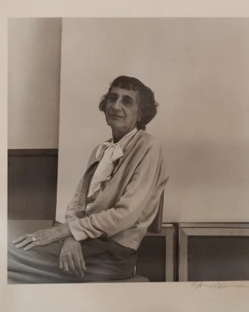 Portrait of an elderly woman with short, dark hair, wearing a light cardigan and scarf, seated indoors against a neutral background.