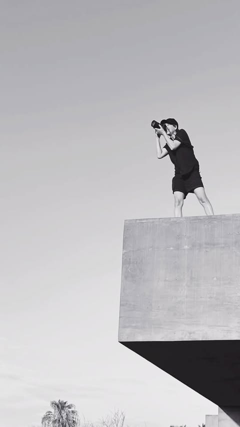 A photographer stands on a concrete structure against a bright sky, holding a camera, wearing shorts, in a black and white, outdoor photo.