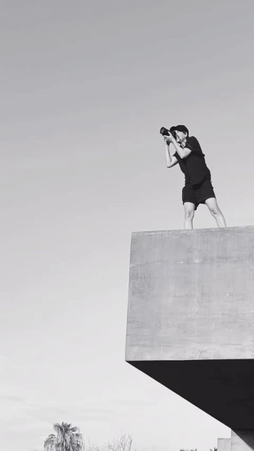 A photographer stands on a concrete structure against a bright sky, holding a camera, wearing shorts, in a black and white, outdoor photo.