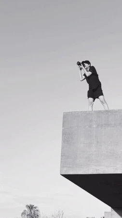 A photographer stands on a concrete structure against a bright sky, holding a camera, wearing shorts, in a black and white, outdoor photo.