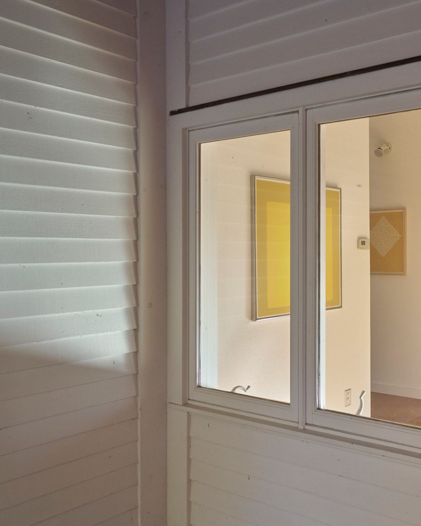 Interior view featuring a white-framed window, shuttered wall, and framed art, capturing a blend of architectural elements and artistic display.